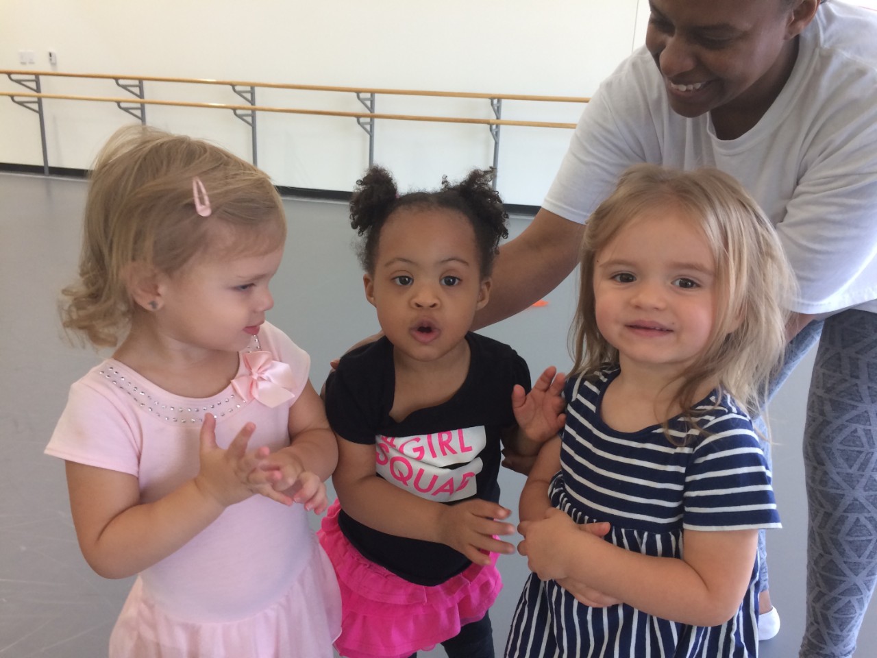 three very young girls in their dance attire. The girl in the middle is a young girl with Down syndrome.