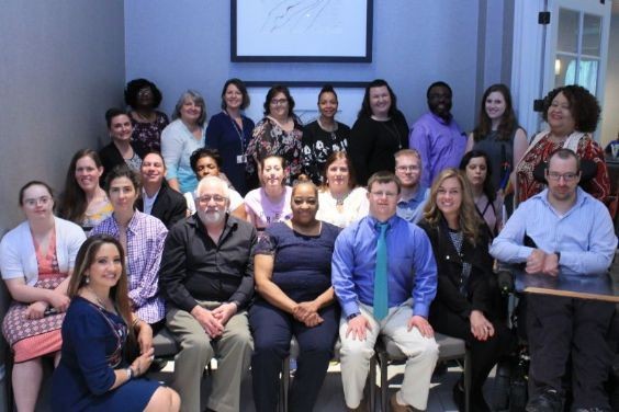 group photo of a diverse group of about 30 folks with and without different types of disabilities, posing outside a conference room in a sunlit hallway
